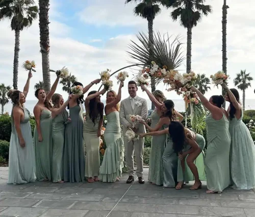 Bridesmaids in sage green dresses forming a bouquet arch over the groom near palm trees in Santa Barbara.