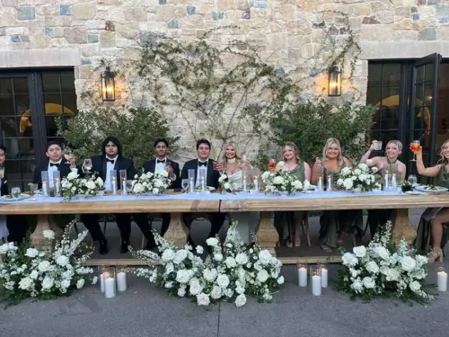 Bride and groom celebrating with their wedding party at an elegantly decorated head table with white floral arrangements during an outdoor wedding reception in Santa Barbara, California.