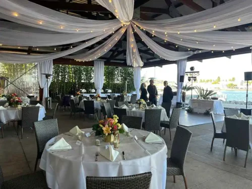 Elegant waterfront wedding reception with white draped ceiling, string lights, round guest tables, and floral centerpieces at a luxury venue in Huntington Beach, California.